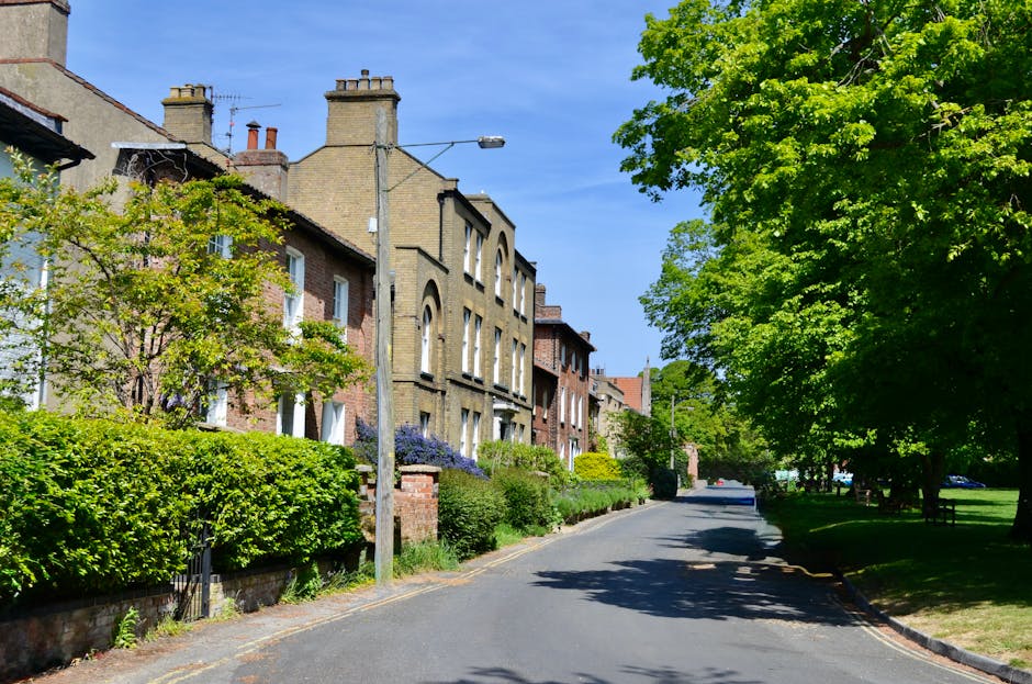 A quiet residential street with a row of Victorian-style terraced houses, each featuring brick facades and chimney stacks, situated alongside a neatly maintained front garden area. The gardens display